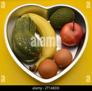 Ein gesundes Herz, eine Auswahl an Obst-, Gemüse- und Milchprodukten in einer herzförmigen Backform vor gelbem Hintergrund. Stockfoto