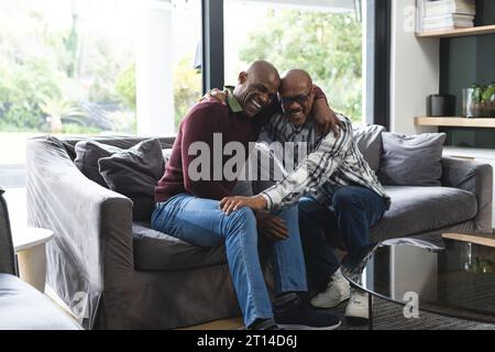 Glücklicher afroamerikanischer Seniorenvater und erwachsener Sohn sitzen zusammen auf der Couch lachend und umarmen Stockfoto