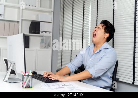 Junger erwachsener asiatischer Geschäftsmann, der auf seinem Schreibtisch mit einem Computer im Hintergrund des Büros gähnt. - Workaholic, langweilig, erschöpft, schläfriges Geschäftsmann-Konzept Stockfoto