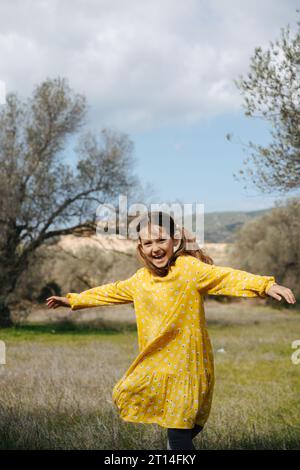 Ein freudiges 8-jähriges Mädchen in einem gelben Kleid dreht sich auf einem Feld vor dem Hintergrund eines Olivenhains. Stockfoto