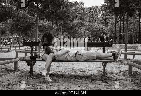 Ein junger Mann liegt auf einer Bank und legt seinen Kopf auf den Schoß seiner Freundin, während sie auf ein Handy im Jardin du Luxembourg, Paris, Frankreich, schaut. Stockfoto