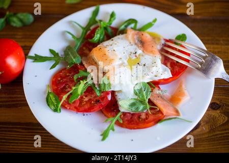 Gekochtes pochiertes Ei mit Kräutern, leicht gesalzenem Lachs und Tomaten mit Gewürzen auf einem Holztisch. Stockfoto