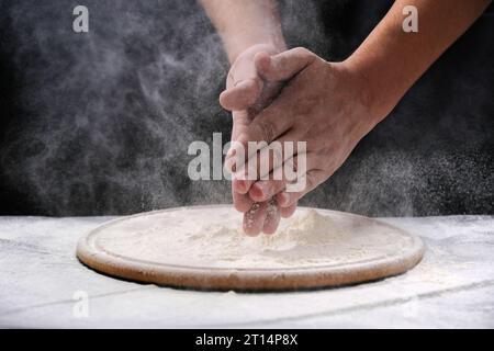 Ein Mann klatscht mit einem Spritzer weißem Mehl und schwarzem Hintergrund in die Hände. Mahlzeit zubereiten, Hände in Mehl Stockfoto