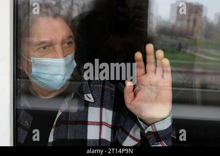 Selbstisolierung Quarantäne. Ein einsamer älterer Mann, der eine medizinische Gesichtsmaske trägt, schaut zu Hause durch das Fenster. Mann in Quarantäne drinnen, Lebensstil Stockfoto