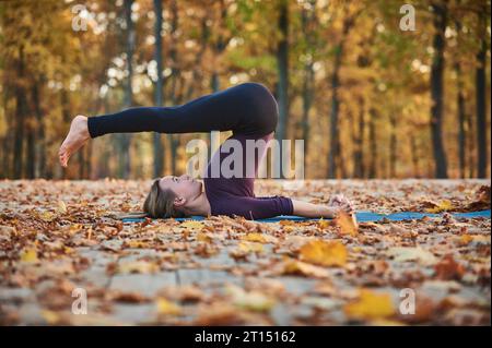 Die schöne junge Frau übt Yoga Asana Halasana Pflug posiert auf der Holzterrasse im Herbstpark Stockfoto