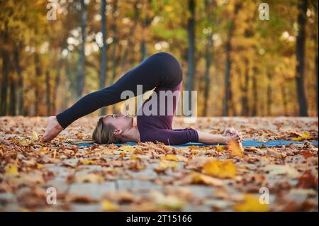 Die schöne junge Frau übt Yoga Asana Halasana Pflug posiert auf der Holzterrasse im Herbstpark Stockfoto