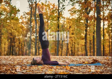 Schöne junge Frau übt Yoga Asana Niralamba Sarvangasana - unbestützte Schulterstütze posiert auf der Holzterrasse im Herbstpark Stockfoto