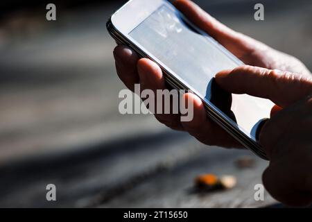 In einem Park ruhen die Hände eines älteren Menschen auf rustikalem Holz und kämpfen mit einem Smartphone. Die Unempfindlichkeit der Technik gegenüber alternden Händen führt zu gesellschaftlicher Ausgrenzung Stockfoto