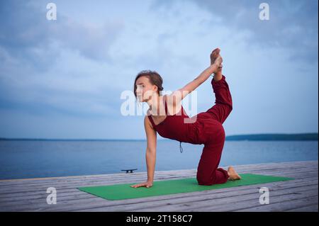 Die schöne junge Frau übt Yoga auf der Holzterrasse in der Nähe des Sees. Stockfoto