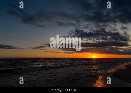 Orangefarbener Sonnenuntergang über dem Meer. Wellen krachen auf den Strand, dramatischer Himmel. Eine statische Aufnahme am frühen Abend Stockfoto