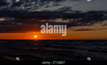 Orangefarbener Sonnenuntergang über dem Meer. Wellen krachen auf den Strand, dramatischer Himmel. Eine statische Aufnahme am frühen Abend Stockfoto