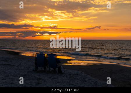 Ältere Ehepaare, die sich am Strand während des wunderschönen Sonnenuntergangs ausruhen. Dramatischer orangefarbener Himmel mit Wolken, ruhige Wellen auf dem Meer. Stockfoto