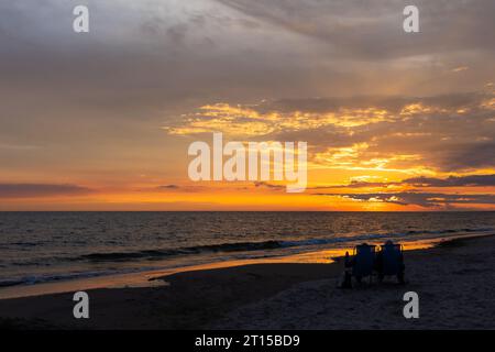 Ältere Ehepaare, die sich am Strand während des wunderschönen Sonnenuntergangs ausruhen. Dramatischer orangefarbener Himmel mit Wolken, ruhige Wellen auf dem Meer. Stockfoto
