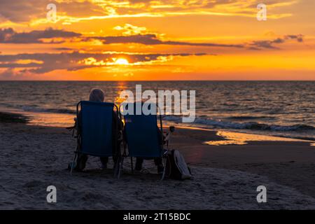 Ältere Ehepaare, die sich am Strand während des wunderschönen Sonnenuntergangs ausruhen. Dramatischer orangefarbener Himmel mit Wolken, ruhige Wellen auf dem Meer. Stockfoto