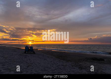 Ältere Ehepaare, die sich am Strand während des wunderschönen Sonnenuntergangs ausruhen. Dramatischer orangefarbener Himmel mit Wolken, ruhige Wellen auf dem Meer. Stockfoto