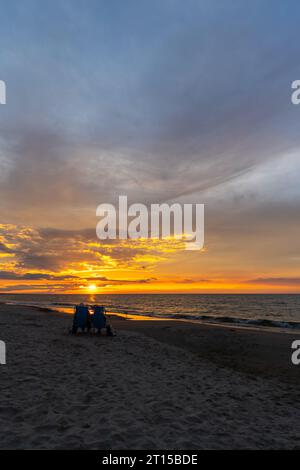Ältere Ehepaare, die sich am Strand während des wunderschönen Sonnenuntergangs ausruhen. Dramatischer orangefarbener Himmel mit Wolken, ruhige Wellen auf dem Meer. Stockfoto