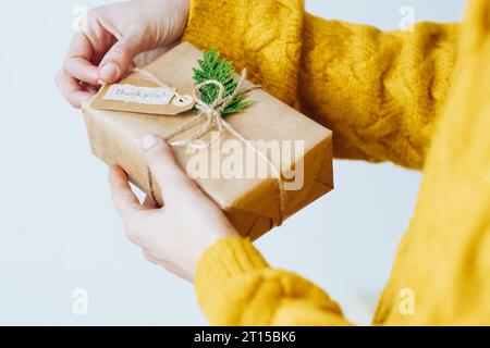 Eine nicht erkennbare Frau in gelbem Pullover mit umweltfreundlicher Geschenkbox mit Etikett mit den Worten Danke. Nachhaltiges und umweltbewusstes Wohnkonzept Stockfoto