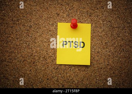 Eine gelbe Papiernote mit der Abkürzung PTSD (posttraumatische Belastungsstörung) darauf, auf eine Korkplatte gesteckt. Nahaufnahme. Stockfoto