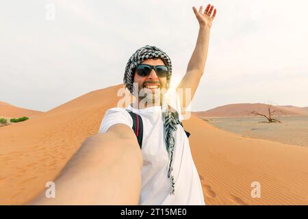 Junger Mann in weißem T-Shirt in der Wüste, auf Safari in den VAE, mit Hut und Rucksack, erkunden die Natur der sandigen Schönheit. Selfie im Rücken machen Stockfoto