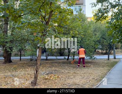 Damenreiniger mit Besenstielbürste reinigt im Herbst Straßen und Rasen von herabfallenden Blättern und Abfällen. Kommunalarbeiter in Uniform mit Besen Stockfoto