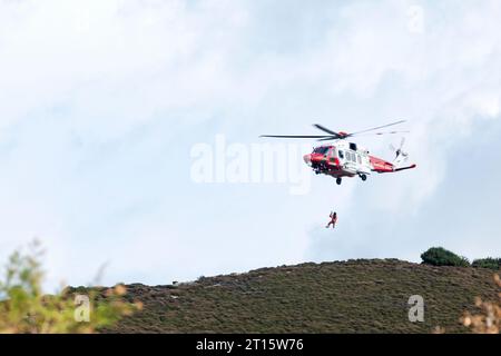 Ein rot-weißer HM Coastguard Hubschrauber, der über einer Landzunge schwebt. Der Hubschrauber windet ein Besatzungsmitglied vom Boden zurück zum Flugzeug Stockfoto