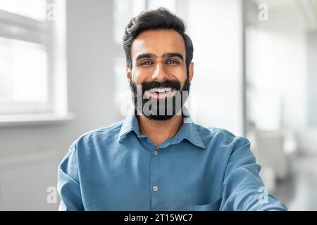 Fröhlich lächelnder indianer, der Hemd trägt, der im Büro sitzt, Nahaufnahme Foto eines glücklichen Geschäftsmannes am Arbeitsplatz Stockfoto