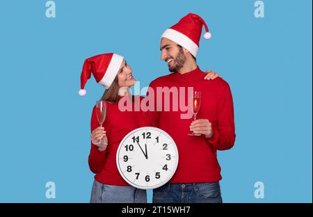 Glückliches Junges Paar In Santa Hats Mit Großer Uhr Und Champagnergläsern Stockfoto