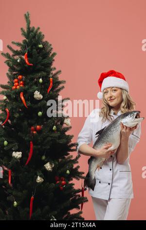 Das Konzept des Kochens und der Weihnachtsferien. Eine schöne Köchin, die einen großen Fisch in der Hand hält, vor dem Hintergrund eines Weihnachtsbaumes. Stockfoto