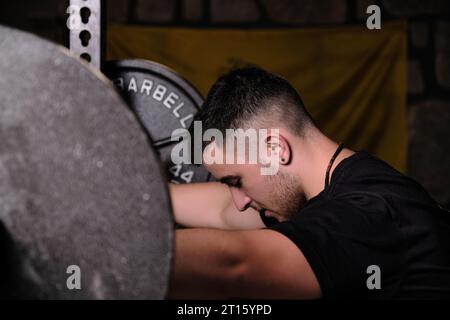 Junger nachdenklicher bärtiger Mann mit Langhantel im Fitnessstudio Stockfoto