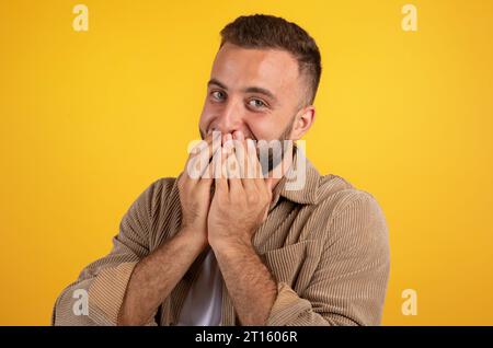 Der lachende, glückliche tausendjährige kaukasier mit Bart in der Freizeit bedeckt den Mund mit Händen, hat Spaß Stockfoto