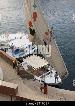 Im Old Cataract Hotel in Assuan blicken Sie hinunter auf eine traditionelle ägyptische Feluke, die neben anderen Segelbooten am Ufer des Nils vertäut ist. Stockfoto