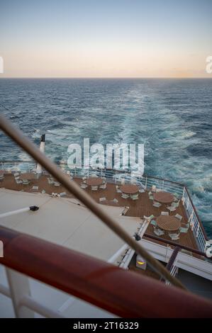 Runde Tische und Stühle stehen auf dem Bootsdeck auf der Rückseite eines Kreuzfahrtschiffes bei Sonnenuntergang, Meer im Hintergrund, vertikale Aufnahme Stockfoto