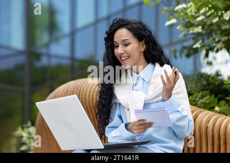 Junge schöne Frau sitzt auf einer Bank vor einem Bürogebäude, benutzt einen Laptop, um ein Webinar-Kursvideo anzusehen, ein Schüler schreibt Daten in ein Notebook, Videoanruf mit einem Lehrer, Fernunterricht. Stockfoto