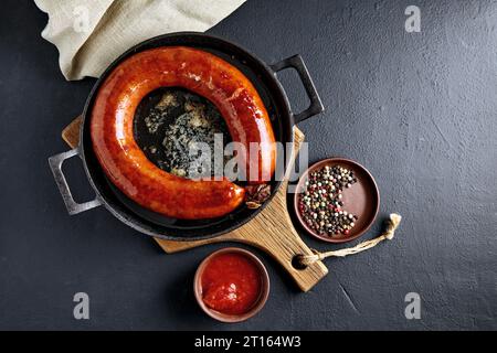 Spiralwurst aus Schweinefleisch, gebraten in einer Pfanne mit kochendem Öl auf einem Holzschneidebrett mit Tomatensauce und Pfefferkörnern auf einem schwarzen Betontisch Stockfoto