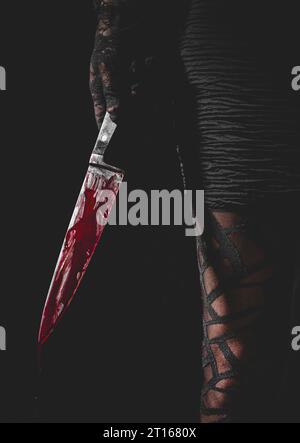 Hand mit schwarzem Spitzenhandschuh hält blutbeflecktes Messer neben dem Bein der Frau, Halloween, dunkler Hintergrund Stockfoto