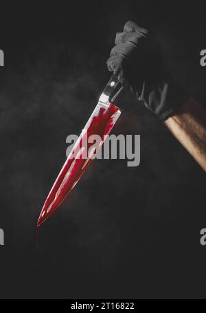 Hand mit schwarzem Handschuh mit blutbeflecktem Messer, Halloween, dunkler Hintergrund Stockfoto
