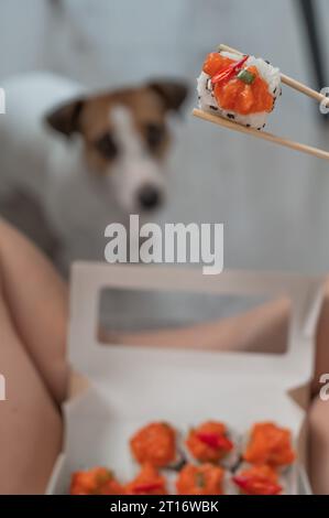 Eine Frau sitzt auf dem Sofa und isst Brötchen. Jack Russell Terrier Hund sitzt auf dem Boden und bittet um Essen von seinem Besitzer. Stockfoto