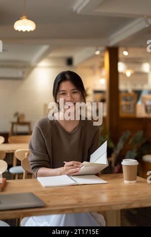 Ein Porträt einer glücklichen und charmanten jungen asiatischen Frau, die fernab in einem schönen Café arbeitet und mit ihrem Buch und ihrer Kaffeetasse an einem Tisch sitzt. Stockfoto