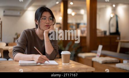 Eine nachdenkliche junge asiatische Frau sitzt an einem Tisch in einem Café, blickt mit einem nachdenklichen Gesicht von der Kamera weg und denkt während der Arbeit an etwas Stockfoto