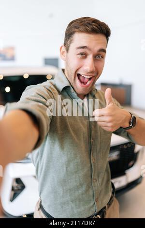 Vertikales POV-Bild eines fröhlichen Käufers, der ein Selfie mit einem Schild auf dem Smartphone vom Händlerbetrieb macht, nachdem er ein neues Auto gekauft hat. Stockfoto