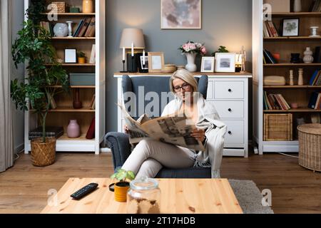 Eine ältere Frau sitzt an ihrem bequemen Sessel in ihrer Wohnung und liest Zeitung, die den Nachrichten folgt. Ältere Frau im Ruhestand liest Informationen über alten sch Stockfoto