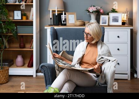 Eine ältere Frau sitzt an ihrem bequemen Sessel in ihrer Wohnung und liest Zeitung, die den Nachrichten folgt. Ältere Frau im Ruhestand liest Informationen über alten sch Stockfoto