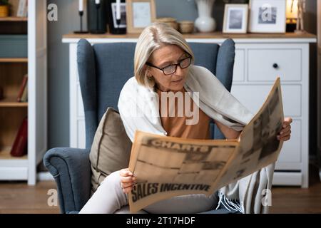 Eine ältere Frau sitzt an ihrem bequemen Sessel in ihrer Wohnung und liest Zeitung, die den Nachrichten folgt. Ältere Frau im Ruhestand liest Informationen über alten sch Stockfoto