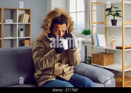 Der ängstliche junge Mann spricht am Telefon und trinkt heißes Getränk, sitzt zu Hause in Strickhandschuhen und warmer Jacke mit Kapuze, friert vor Kälte Stockfoto