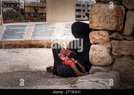 Eine arme muslimische Frau in einem schwarzen Hijab sitzt auf dem Boden und hält ihr Baby in den Armen vor dem Hintergrund einer Stadt im Nahen Osten. Konzept von p Stockfoto
