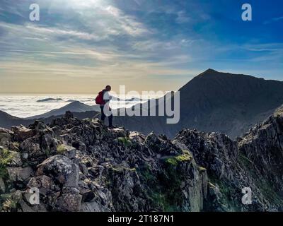 Einsamer Kletterer auf Crib Goch mit CRI-y-ddysgl und Mount Snowdon in der Ferne, mit niedrigen Wolken, die das Tal dahinter füllen. Wales. UK Stockfoto
