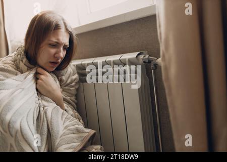 Eine leidende Frau im Karo sitzt an der Heizbatterie, wärmt sich im Haus auf und friert ein. Stockfoto