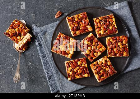 Hausgemachte Toffee gemischte Nüsse Shortbread Bars auf schwarzem Teller auf grauem Betontisch mit Kuchenschaufel, horizontale Ansicht von oben, flache Lage Stockfoto