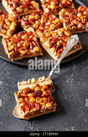 Toffee gemischte Nüsse Shortbread Bars auf schwarzem Teller und ein Stück auf Kuchenschaufel auf grauem Betontisch, vertikale Ansicht, Nahaufnahme Stockfoto