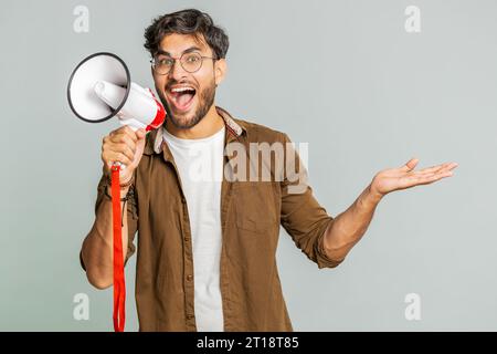 Indischer junger Mann, der mit Megaphon spricht, Nachrichten verkündet, Werbung Rabatte verkauft, Lautsprecher benutzt, um Sprache zu schreien. Glücklicher Araber, isoliert auf grauem Hintergrund Stockfoto